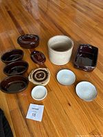 Photo showing 11 vintage pottery pieces in brown and white colors arranged on wood floor. Includes multiple serving bowls, lidded container, crock jar, trivet, and ceramic dishes.
