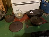 Photo showing vintage cast iron kettle, aluminum pot, large rectangular Dutch oven, round cast iron pot, and two lids on green floor in front of a white oven.