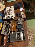 Photo showing vintage cassette and microcassette recorders, multiple walkie talkies, portable radio, backup camera system box, and small cassette tapes on wood table.