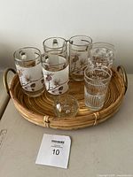 Top view of eight pieces of vintage glassware arranged on wicker tray including mugs with pinecone motif, etched glasses, clear tumbler, wicker tray and glass paperweight on table.