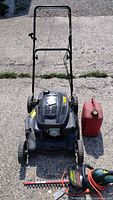 Front view of Yardworks gasoline lawn mower, red gasoline container, and electric hedge trimmer on concrete surface.