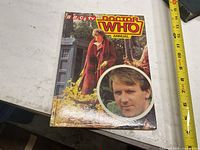 Front cover of the Doctor Who Annual book showing character in plaid coat and a circular inset image of a manâ€™s face.