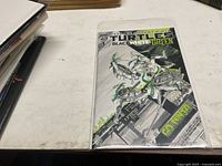 Photo of Ninja Turtles comic book titled 'Black White Green' issue 1, protected in plastic sleeve and placed on table with other papers nearby.