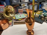 Copper pitcher placed on a white table surrounded by various household items including a brass horn, copper pot, books, and paper.