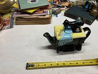 Photo shows ceramic teapot shaped like an old sewing machine with a measuring tape next to it showing about 7.5 inches in length.