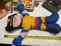 Front side of Wolverine plush doll lying on table with measuring tape underneath indicating about 18 inches in length.