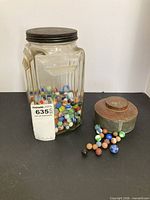 Jar partially filled with assorted marbles beside metal tin lid and loose marbles