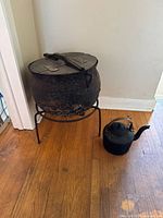 Antique cast iron pot on metal stand with wood lid, next to a cast iron kettle on wooden floor.