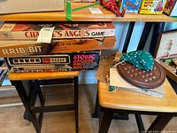 Vintage board and tabletop games stacked on a table, showing box labels for Charlie's Angels, Rib-bit, and Atomic Arcade Pinball games, plus a round wooden marble solitaire board.
