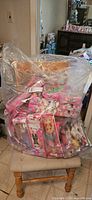 Large clear plastic bag filled with dolls in pink boxes on a chair.