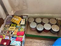 Box containing various packaged soaps including Camay, cocoa butter, baby soap, goat's milk soap and more on left and a box of eight White Barn candles in decorative tins on right