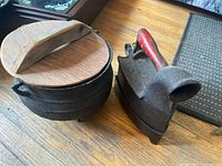 Photo showing both the cast iron cauldron with wooden lid and the vintage coal iron with red handle on a wooden floor.