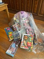 Photo showing multiple sealed toys including doll in ice cream shape, toy food playsets with burger and fries, and other boxed toys in a plastic bag on wooden floor.