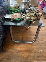 Photo showing the smoked glass top and chrome tubular metal base of the side table, with decorative metal items placed on top (items not included).