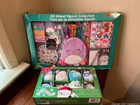 Wide shot of All About Squish Collection with multiple stationery items and a small purple plush pouch in original packaging, with a boxed set of 8 Squishmallow ornaments beneath it.