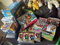 Photo of a variety of toys including Giggle Bots, Tenkai Knights, Transformers and others, many still sealed in boxes or blister packs, laid out on a couch with some plush toys in the background.