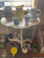Full view of the demi-lune table topped with various pottery vases and jugs, some pottery pieces below on the shelf and surrounding floor