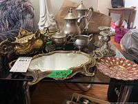 Full set displayed on a black glass table showing silver and brass tea and coffee pots, cream and sugar containers, ornate mirrored tray, brass creamer or pitcher, and a small brass decorative piece