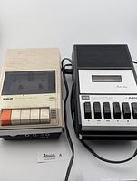 Two vintage cassette recorders side by side: left is off-white RCA unit with orange and white buttons; right is black and silver Aiko recorder with black buttons and label