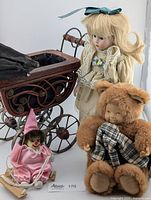 View of the porcelain doll next to the antique baby carriage, with the Anne Geddes Baby Bear Doll in front and the hanging clown figurine positioned on the left