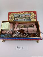 Open decorative footed tin box with scenic painted lid showing multiple jewelry pieces including necklaces, bracelets, eyeglasses, and small boxes inside.