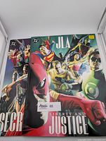 Both oversized JLA graphic novels side by side, clearly showing titles "Liberty and Justice" and "Secret Origins," with several Justice League heroes depicted on the covers.