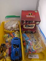 Yellow bin containing a blue LEGO car, plastic zip bags with LEGO pieces and figures, an Incredibles 2 Glow Buddy boxed figure