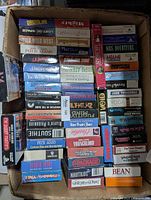 Top-down view of many VHS tapes in original boxes stacked inside a cardboard box showing various titles including Patch Adams, Mrs. Doubtfire, Nautilus, Wild Wild West, final voyage and others.