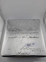 Embossed silver metal storage case with Monopoly logo and Mr. Monopoly character, showing surface wear and damage on edges