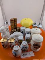 Various salt and pepper shakers including ceramic bunnies, floral gold porcelain, wooden, crystal, mushroom-shaped, and souvenir styles, shown on an orange tray.