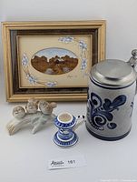 Photo showing all items together: framed art, Delft blue candlestick holder, porcelain bird figurine on a branch, and Handarbeit stein with pewter lid.