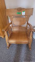 Front view of the light brown wooden rocking chair showing carved wood details in Victorian style on the top backrest panel, seat, and arms.