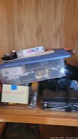 Shelving with plastic bin containing assorted small electronics and accessories alongside a black electronic device possibly a cassette or radio player.