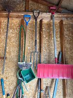 Photo showing multiple garden and yard tools hanging on a wooden wall including rakes, shovels, forks, a green hand scoop, and a red snow shovel.
