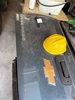 Black Chevrolet Silverado tailgate with bowtie emblem and handle on a workshop surface with yellow hardhat partly visible.