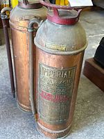 Two vintage copper and brass fire extinguishers with visible labels, patina, and metal tubing. One has a large Imperial soda & acid usage label, other shows side rivets and tubing.