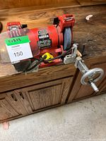 Full view of the Kango bench grinder on a wooden surface showing grinding wheel, tool rest, crank handle, and power cord with yellow switch.