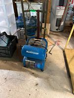 Blue Mastercraft air compressor positioned on concrete floor in basement or workshop setting, showing full side view including handle and wheels.