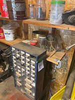 Full view of black parts organizer bin with small drawers on a wooden shelf filled with various jars and containers of hardware items in a workshop.