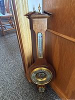 Full view of weather station showing wooden body with brass top finial and mounted barometer, thermometer, and small dial.