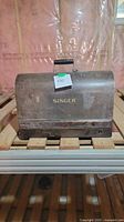 Front view of vintage Singer sewing machine's original hard case showing the worn brown finish with 'SINGER' in gold lettering and a black handle on top.