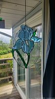 Blue iris flower stained glass suncatcher hanging in window showing translucent blue petals and green leaves within oval clear glass