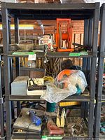 Photo showing three metal shelves holding various workshop tools and parts including orange hose reel, electric tools, metal parts, and containers with visible dust and rust.