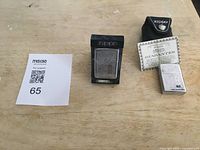 Overview image showing two Zippo lighters side by side on a wooden surface with a maxsold lot card. One lighter is upright in a black plastic Zippo stand. The other lighter is next to a black leather Zippo pouch and a Zippo guarantee card.