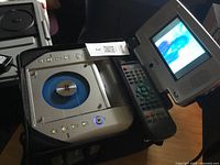 Audiovox portable DVD and VHS player side view showing the remote control, disc loading area with blue disc clamp, control buttons, and flip screen displaying video.