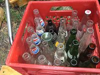 Red plastic crate filled with multiple vintage glass soda bottles including Pepsi-Cola, Coca-Cola and other brands in clear, green, and brown glass.