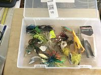Opened clear tackle box with various used fishing lures visible inside including frog lures, spinnerbaits, and soft plastic baits.
