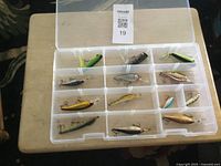 Top-down photo of clear plastic box showing 15 compartments each with a fishing lure. Lures vary in size and color with attached hooks.