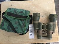 Camouflage binoculars and matching fabric carrying case displayed on light wood tabletop with auction tag. View of binoculars from front and side showing structure and grip.