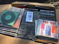 Box filled with approximately 150 used music CDs in jewel cases, some album titles visible like Rush Chronicles and Doo Wop Album.